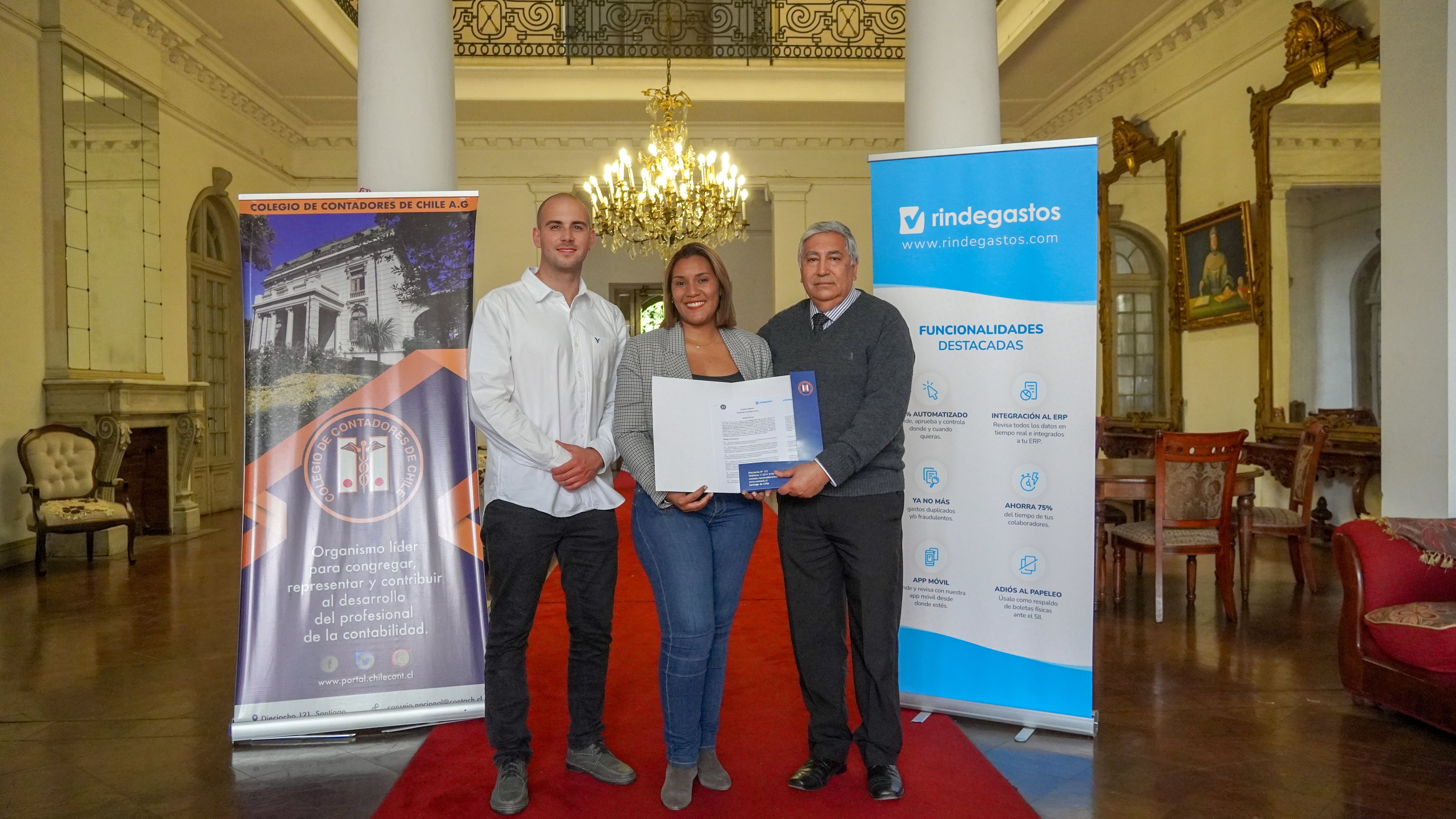 Representantes de Rindegastos y el Colegio de Contadores de Chile posan con un documento de alianza, en un elegante salón con candelabro, destacando la colaboración entre ambas instituciones.