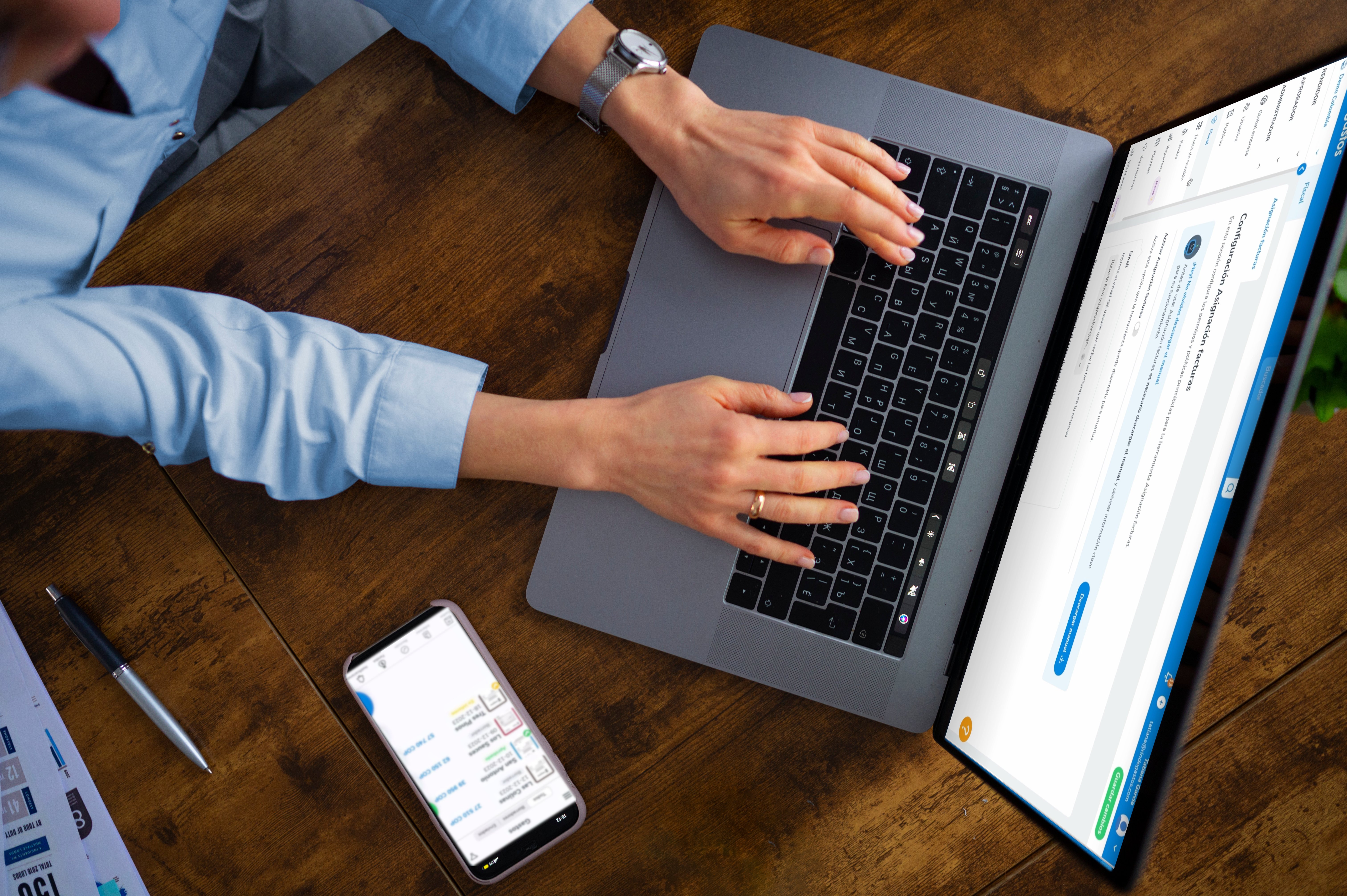 Vista superior de una mujer usando una computadora portátil sobre un escritorio de madera. La pantalla de la computadora muestra una interfaz de software con una sección titulada 'Contabilización Automática Financiera'. Al lado del portátil, hay un teléfono móvil mostrando una aplicación con varias opciones y un bolígrafo junto a un documento en el escritorio.