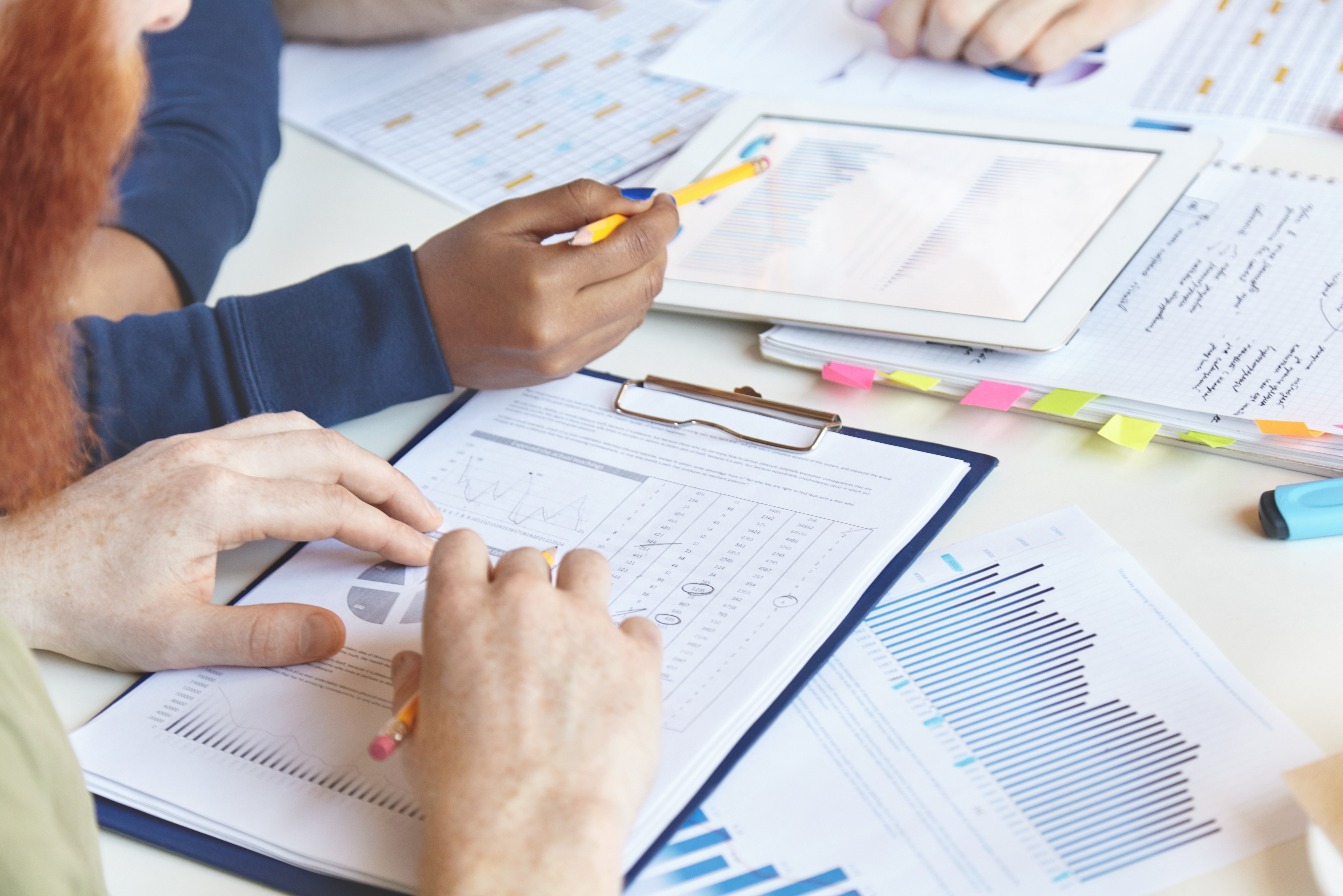 Varias personas colaborando en un análisis de datos y gráficos en una mesa, con hojas impresas y una tablet entre los materiales de trabajo.