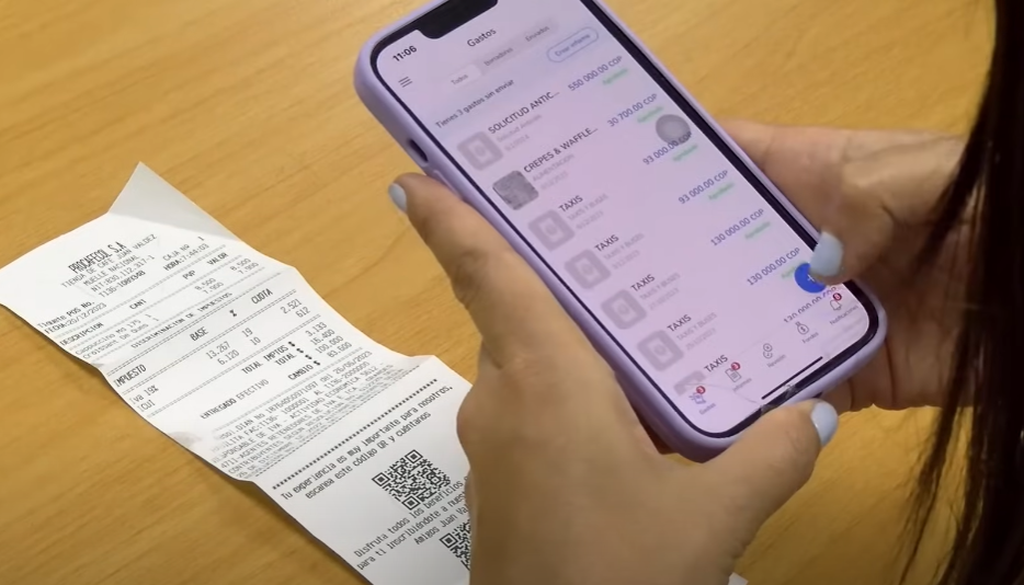Manos de una persona sosteniendo un teléfono móvil con la aplicación de Rindegastos abierta, mostrando una lista de gastos. Junto al teléfono, sobre la mesa de madera, hay un recibo de papel, lo que indica que la persona está digitalizando o verificando gastos físicos utilizando la aplicación.