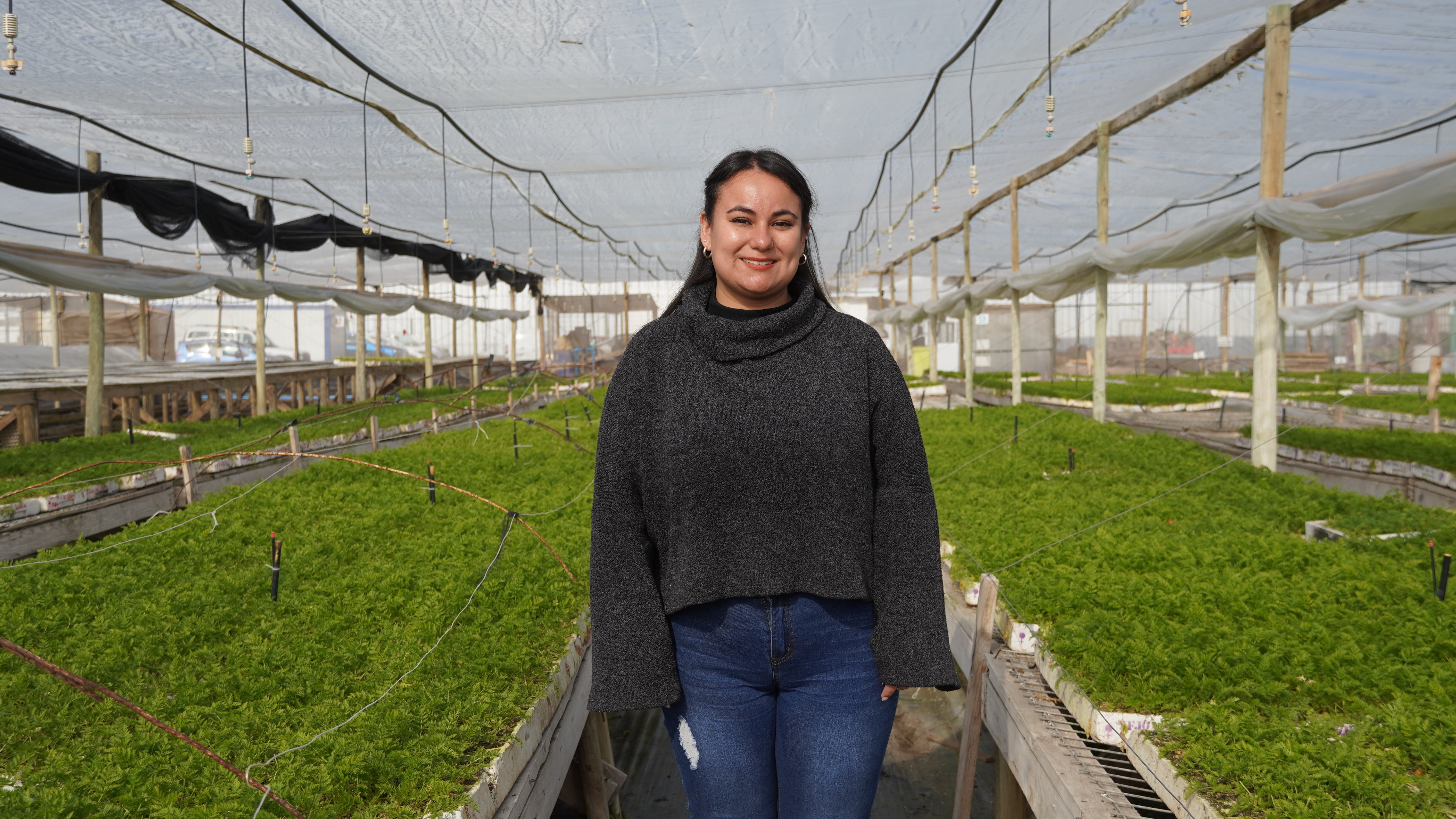 Una mujer con suéter oscuro está de pie en un invernadero de plantas, sonriendo a la cámara. La estructura es similar a la anterior, con filas de plantas verdes extendiéndose a lo largo del espacio.