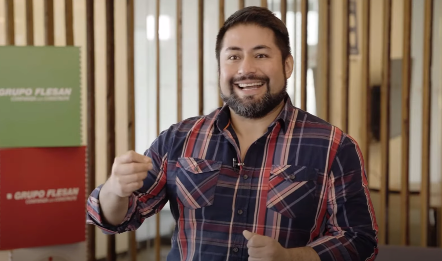 Hombre sonriente con camisa de cuadros hablando frente a la cámara, en una oficina del Grupo Flesan con logotipos de la empresa en el fondo.