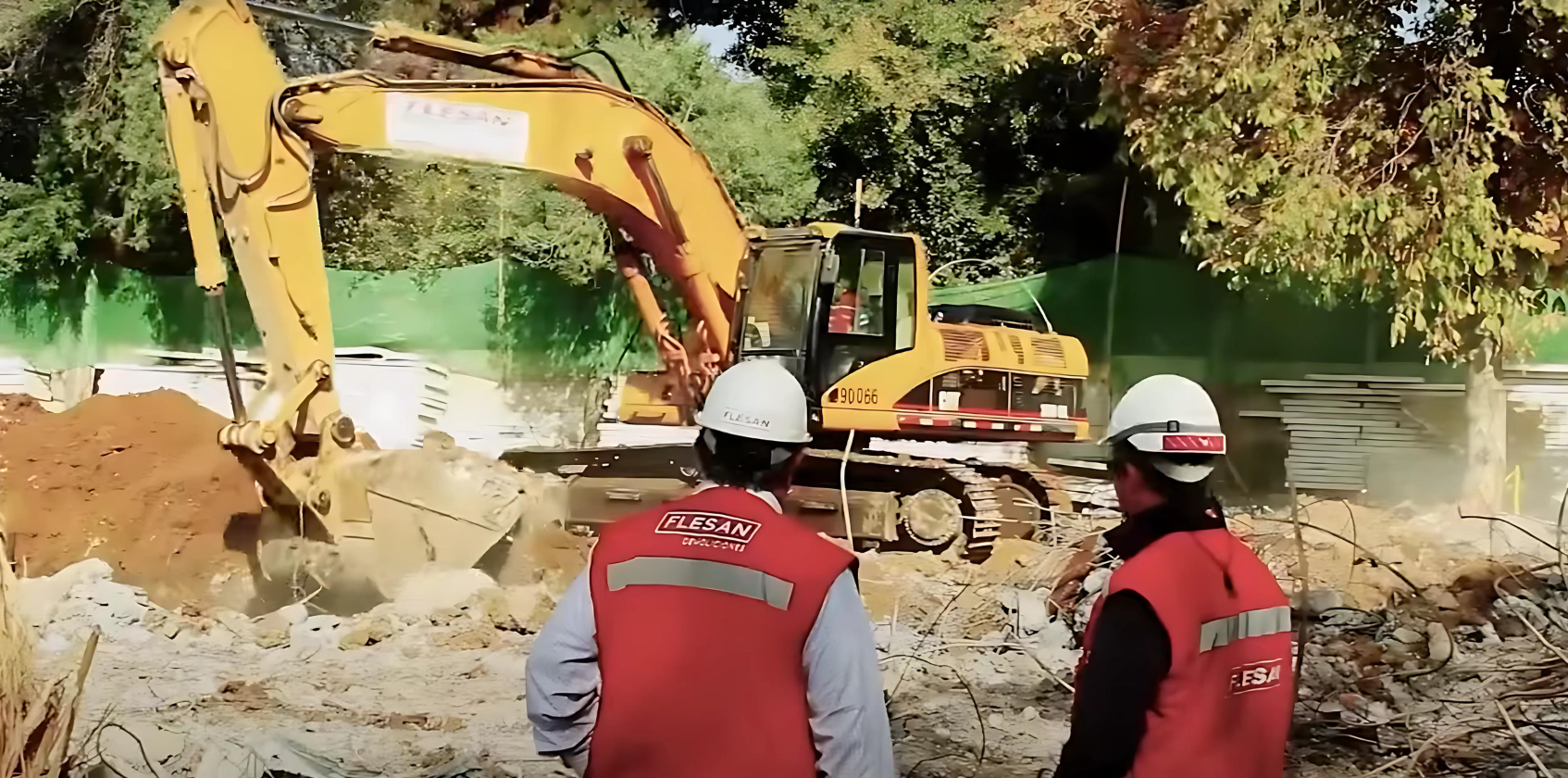 Trabajadores del Grupo Flesan observando una excavadora en acción durante una obra de construcción, ambos con chalecos y cascos de seguridad.