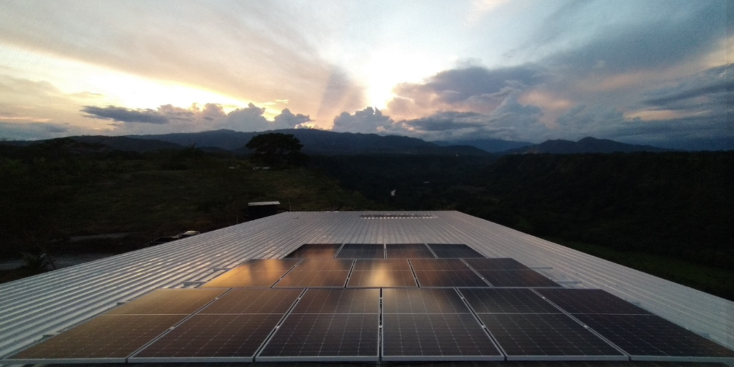 Paneles solares instalados en un techo blanco con un atardecer de fondo, mostrando un paisaje montañoso y nubes iluminadas por la luz del sol.