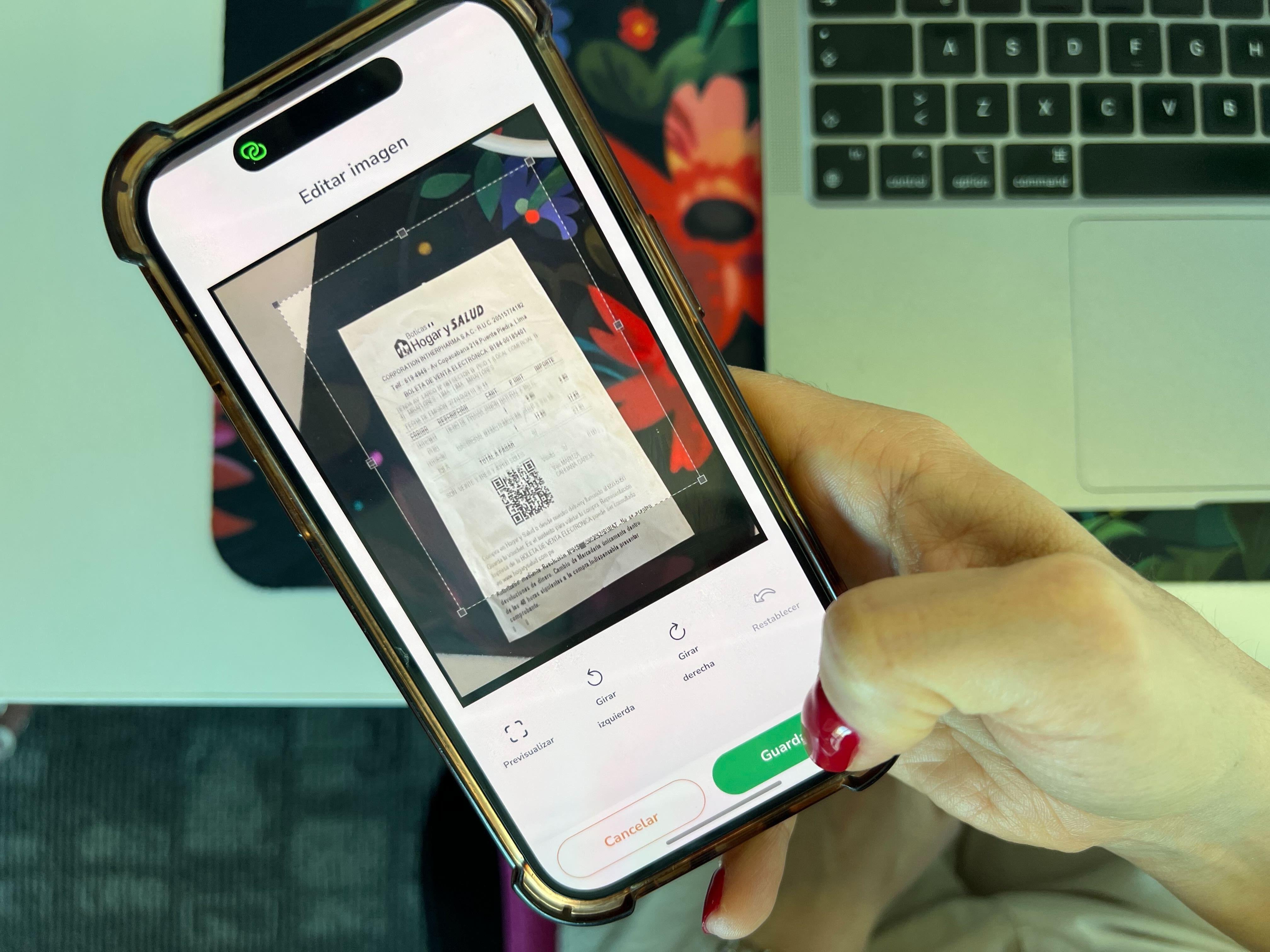 Primer plano de una mano sosteniendo un teléfono móvil que muestra un recibo escaneado en una aplicación, con una computadora portátil en segundo plano."