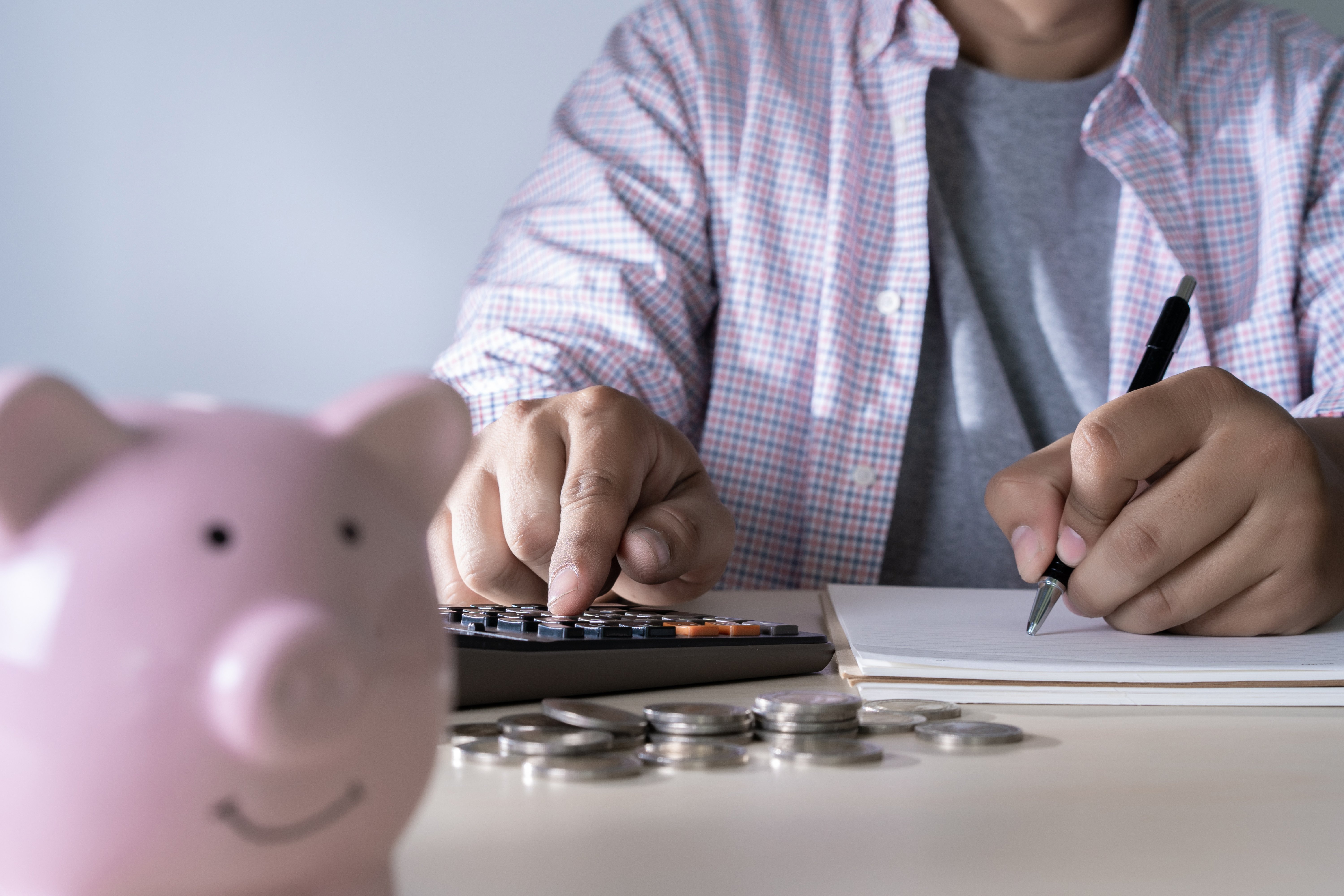 "Primer plano de manos utilizando una calculadora y escribiendo en un cuaderno, con monedas y una alcancía en forma de cerdito en la mesa, simbolizando ahorro y planificación financiera