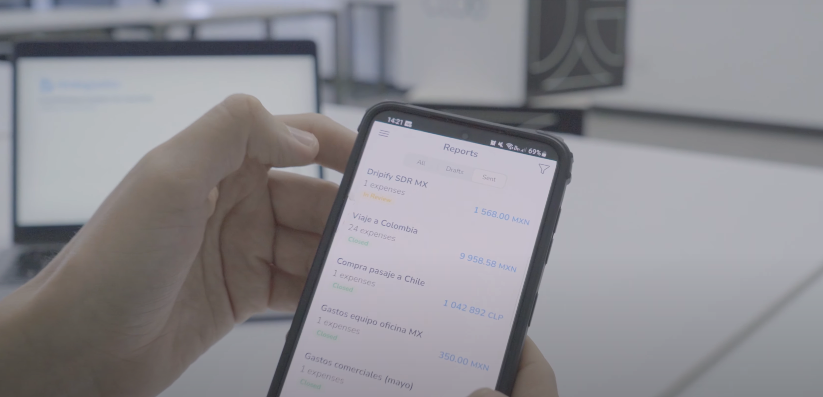 Mano sosteniendo un teléfono móvil que muestra la pantalla de la aplicación de Rindegastos con una lista de reportes de gastos, cada uno con su respectivo monto y estado. El fondo muestra una oficina con una laptop desenfocada, lo que sugiere un entorno de trabajo enfocado en la gestión de gastos y la revisión de informes desde un dispositivo móvil