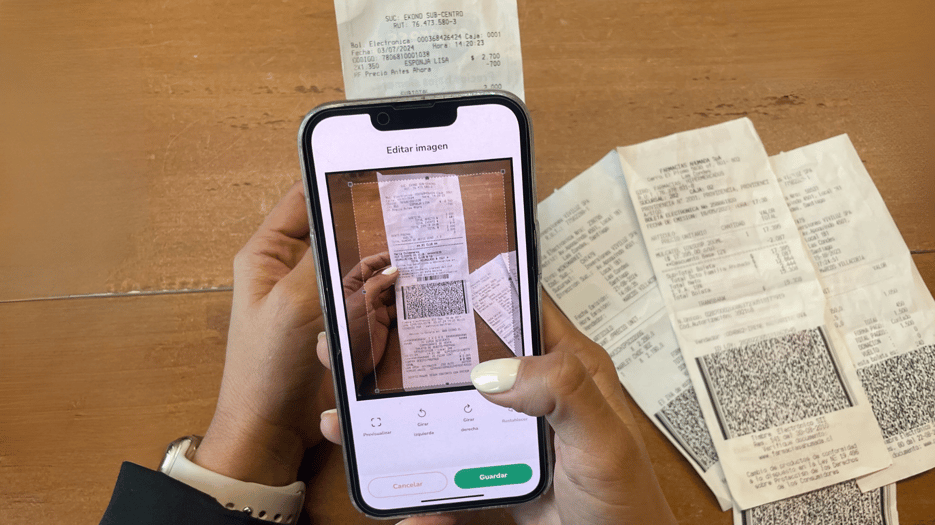 Mano de una persona sosteniendo un smartphone, utilizando una aplicación para tomar una foto de un recibo en papel. La pantalla del teléfono muestra opciones de edición para la imagen capturada, con varios recibos de papel esparcidos sobre una mesa de madera en el fondo.