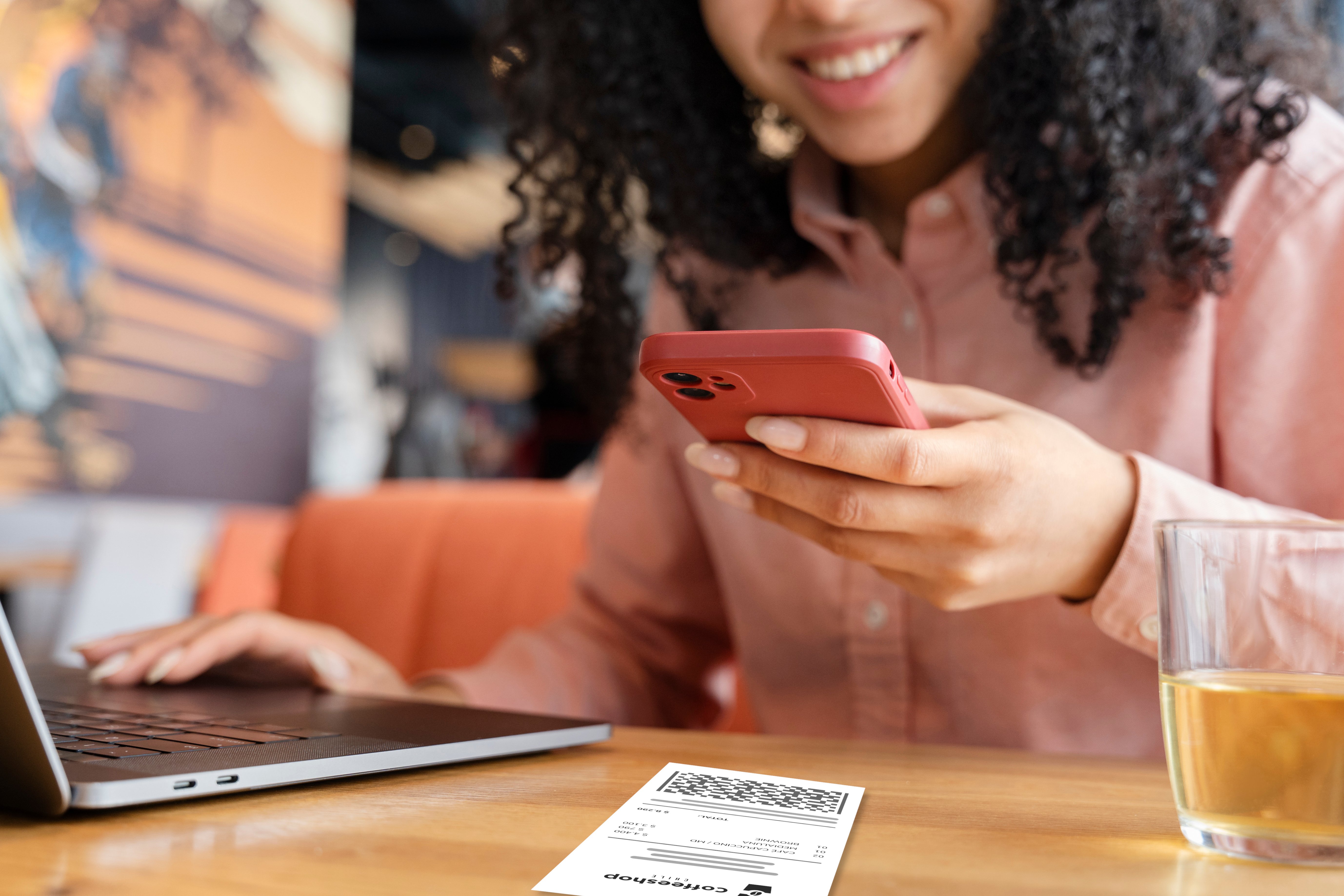 Una persona sostiene un smartphone mientras está sentada en una mesa, capturando una imagen de un recibo. En la mesa hay un laptop abierto y un vaso con bebida al lado.