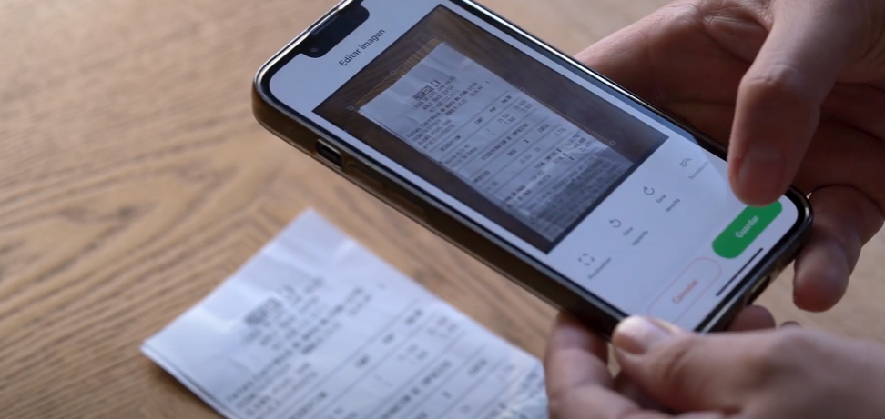 Primer plano de un teléfono móvil escaneando un recibo de gasto sobre una superficie de madera, destacando la tecnología de digitalización de Rindegastos