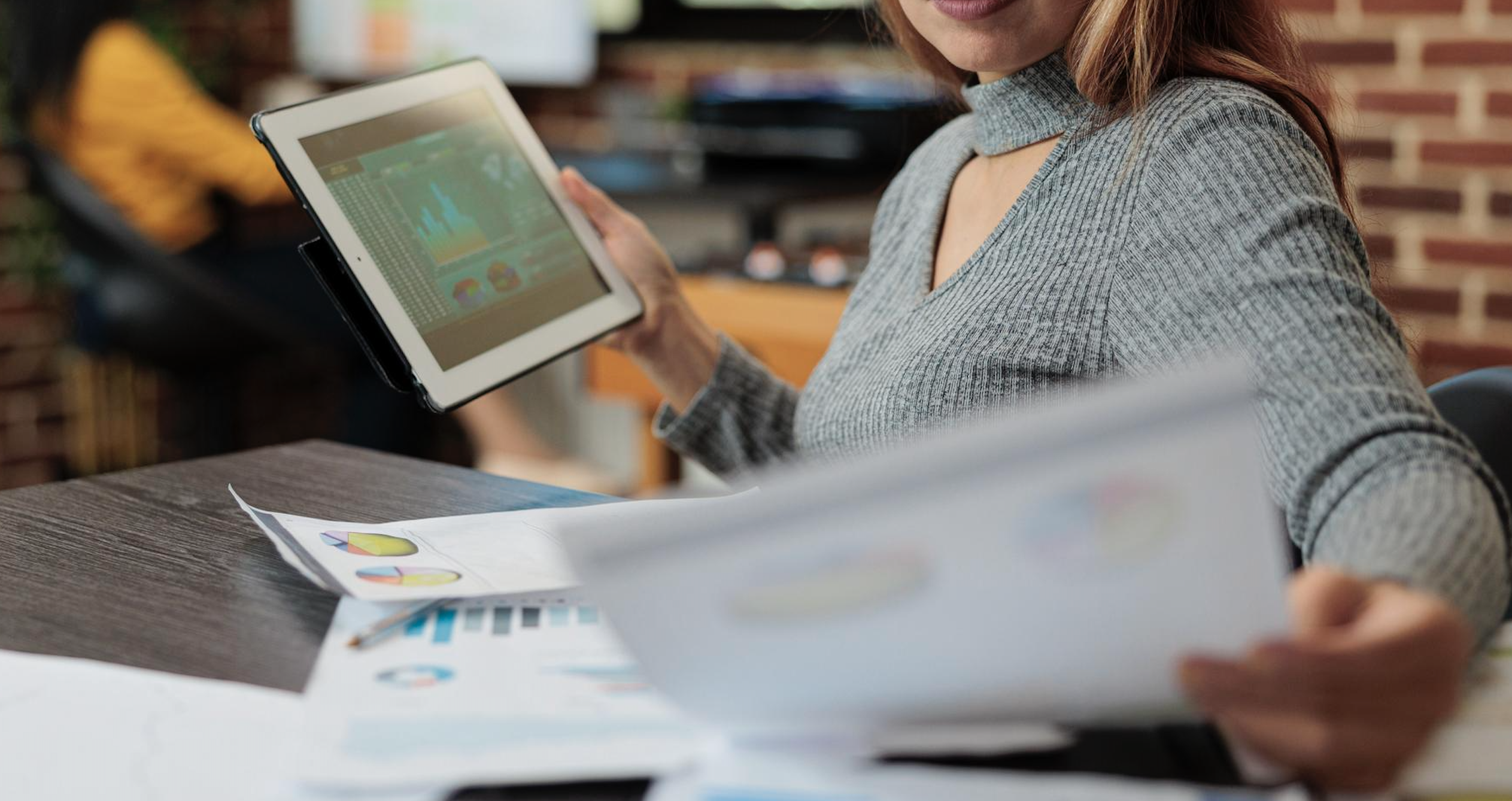 Mujer en oficina revisando reportes financieros en una tablet, con documentos impresos y gráficos en una mesa de trabajo.