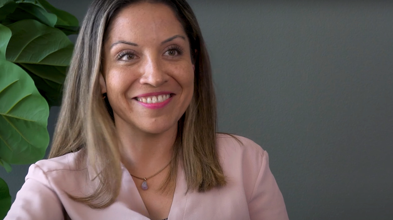 : Primer plano de una mujer sonriente durante una entrevista o conversación en un entorno profesional, con una planta decorativa en el fondo.