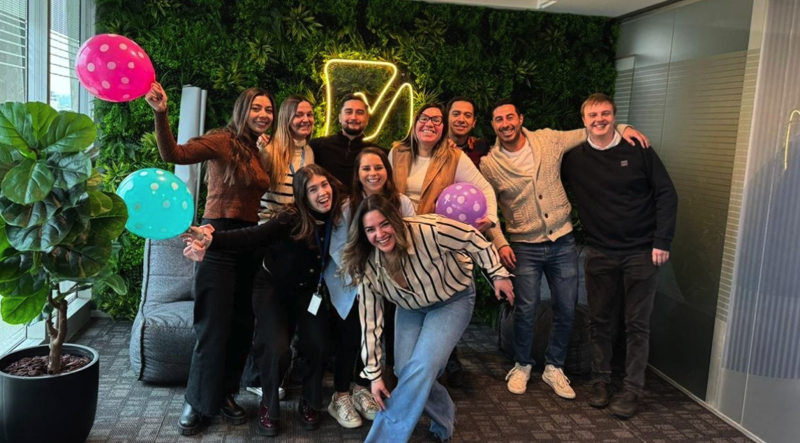  Un grupo de colaboradores de Rindegastos sonrientes reunidos para una foto en una oficina decorada con plantas, sosteniendo globos de colores, en lo que parece ser una celebración o evento informal de equipo.