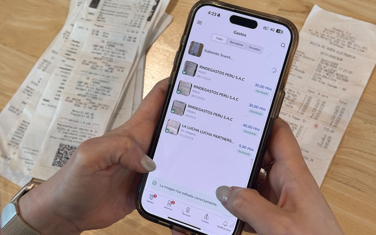 Manos sosteniendo un teléfono móvil mostrando una lista de gastos escaneados en la aplicación Rindegastos con recibos en el fondo.