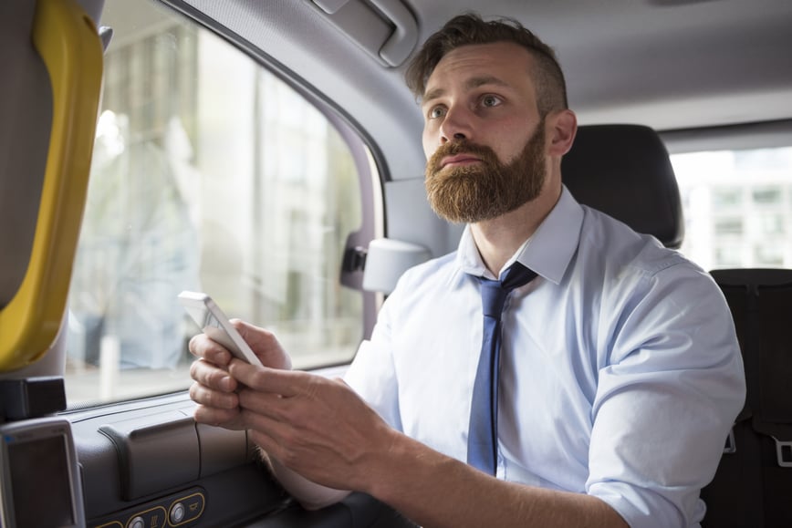 Hombre de negocios con barba utilizando su teléfono móvil en la parte trasera de un coche, enfocando en una aplicación, posiblemente relacionada con la gestión de gastos de viaje