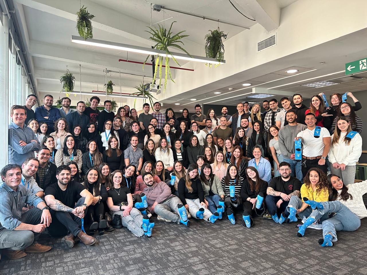 Foto grupal de gran tamaño en la que aparecen decenas de colaboradores de Rindegastos, todos posando sonrientes con calcetines de color azul con el logo de la empresa. Están reunidos en una sala luminosa con plantas colgantes.