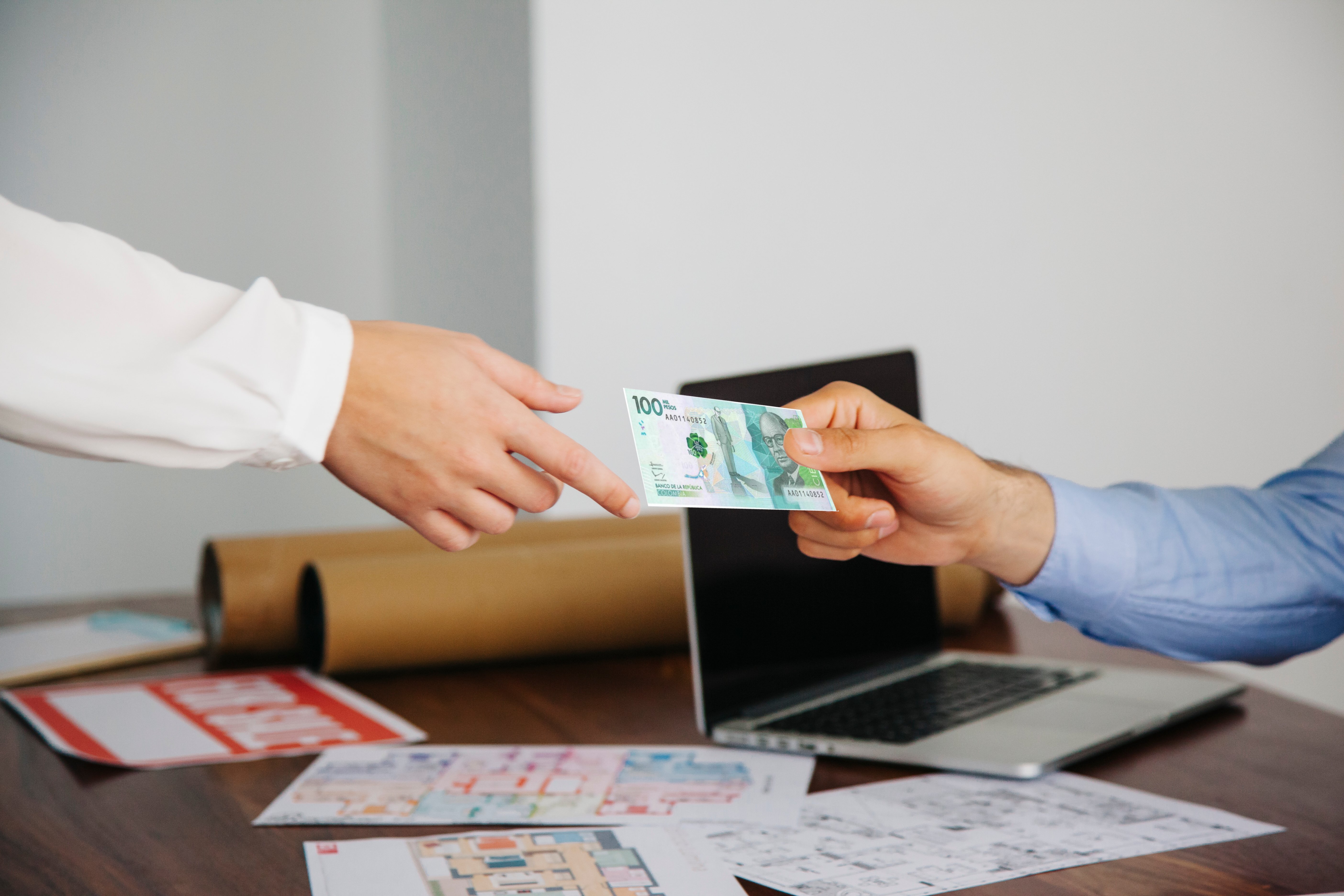 Dos personas realizando una transacción de dinero en efectivo, intercambiando un billete, con planos en la mesa de fondo.