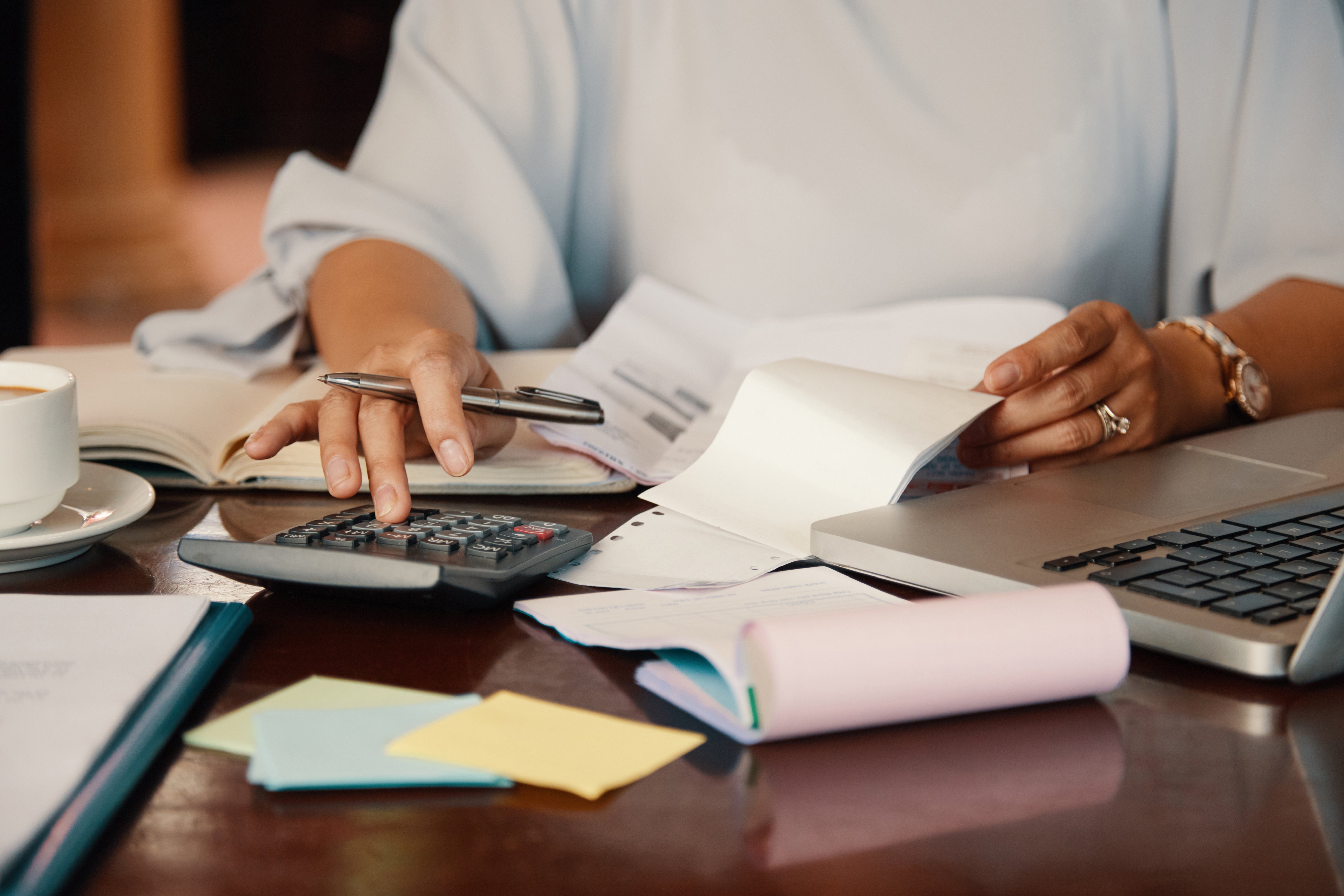 Persona utilizando una calculadora para realizar cálculos con recibos y documentos sobre una mesa, junto a una computadora portátil y una taza de café