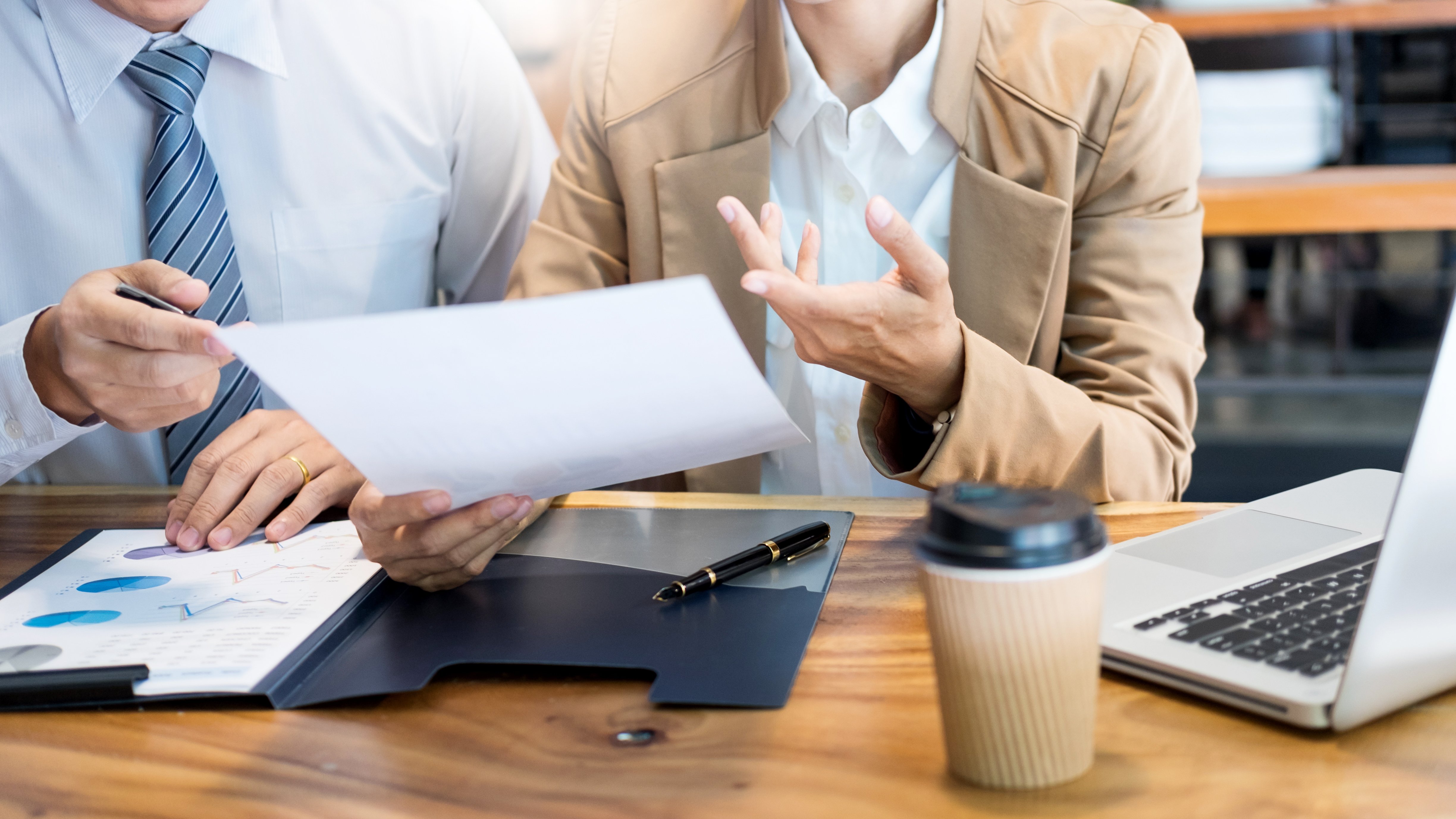 Dos personas revisando documentos financieros sobre una mesa con gráficos y un computador portátil en un entorno de oficina.