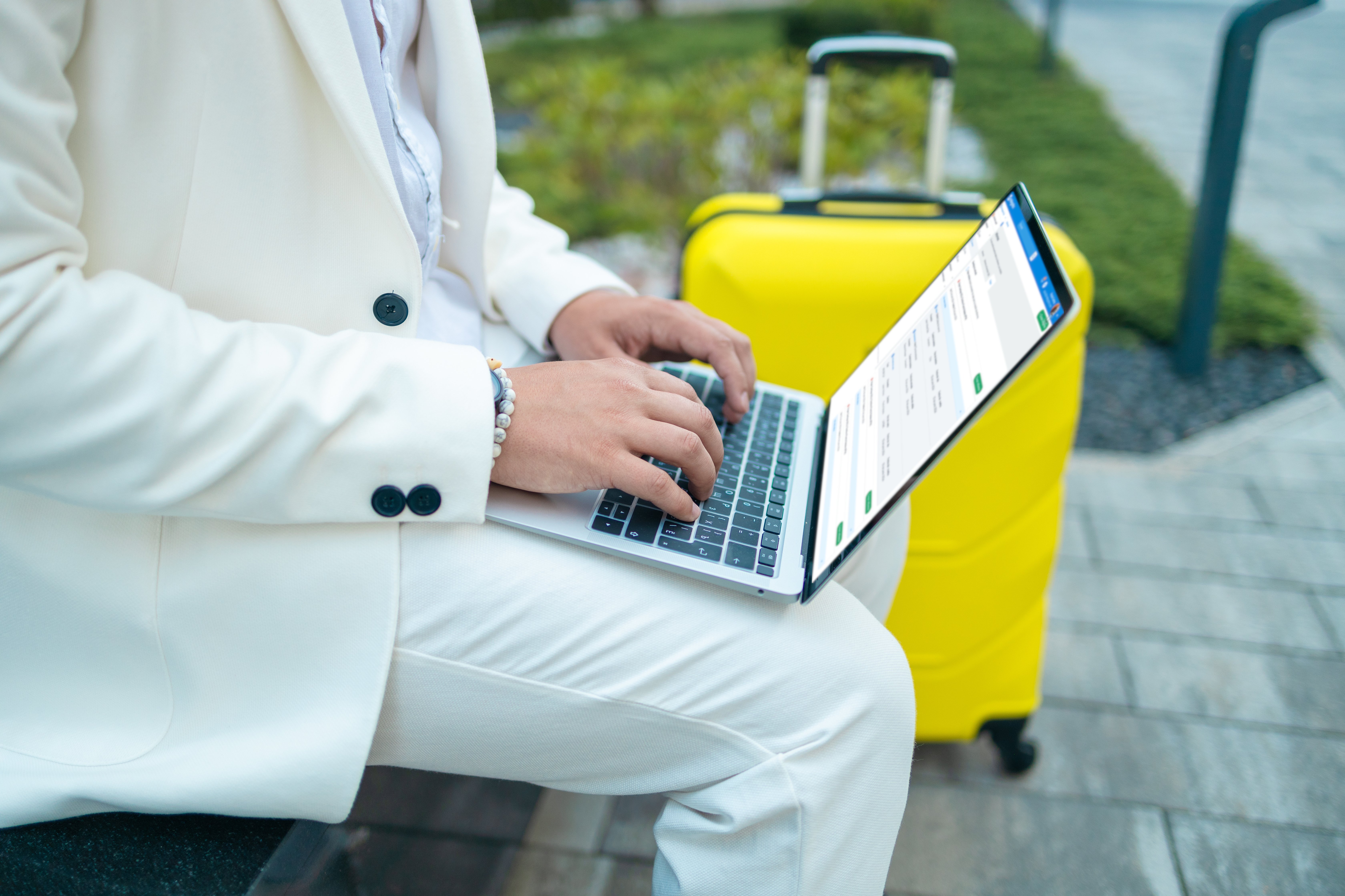 Persona usando una laptop mientras se encuentra al aire libre, con una maleta amarilla junto a ella, visualizando un reporte de viajes en la pantalla.