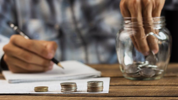 Hombre sacando monedas de un tarro de ahorro mientras toma notas en un cuaderno