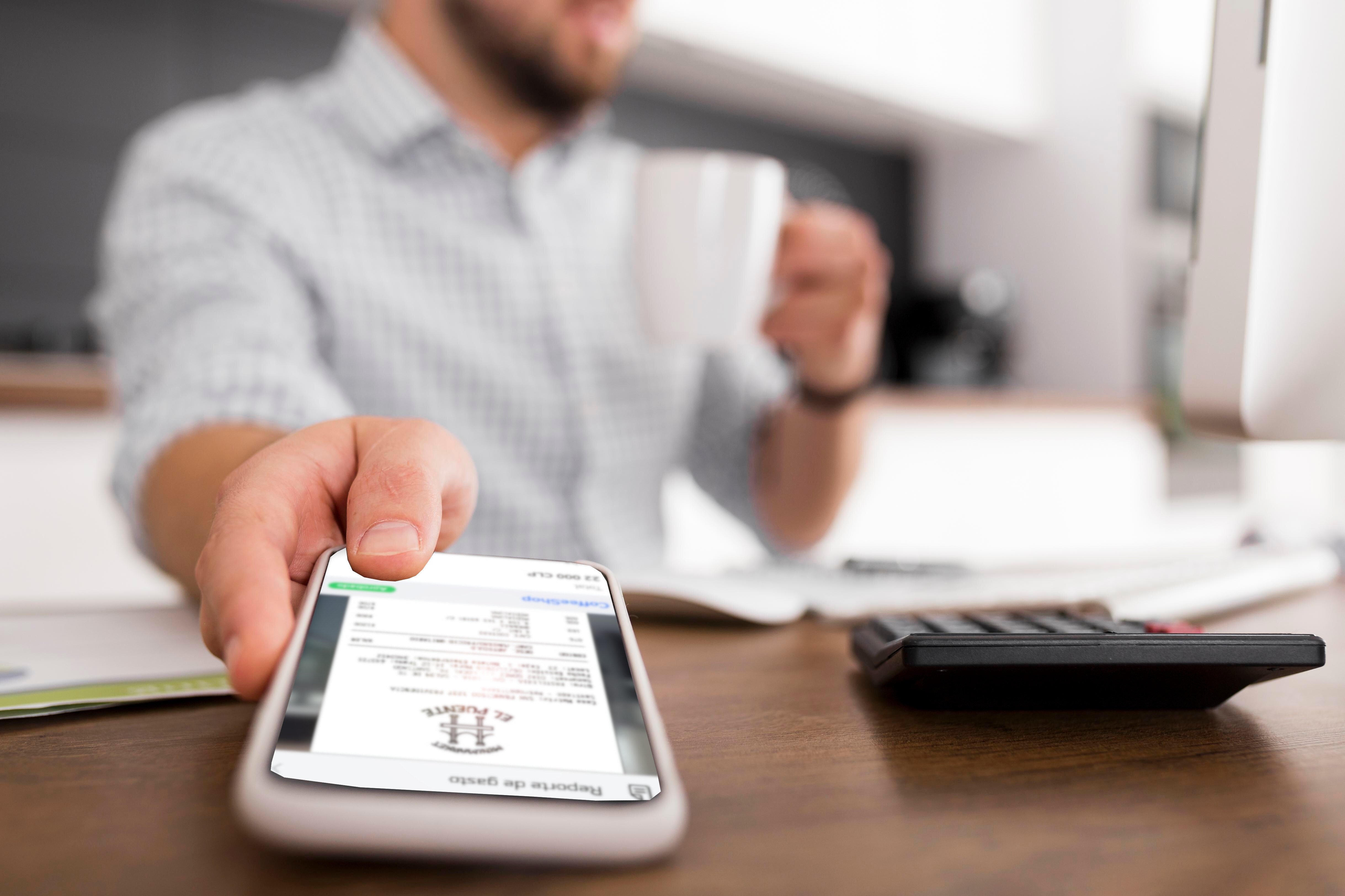 Hombre sosteniendo una taza de café mientras usa un teléfono móvil en una oficina. En la pantalla del teléfono se muestra un recibo digital con detalles de un gasto desde Rindegastos. En el escritorio hay una calculadora y documentos, lo que sugiere un entorno de trabajo enfocado en la gestión financiera.