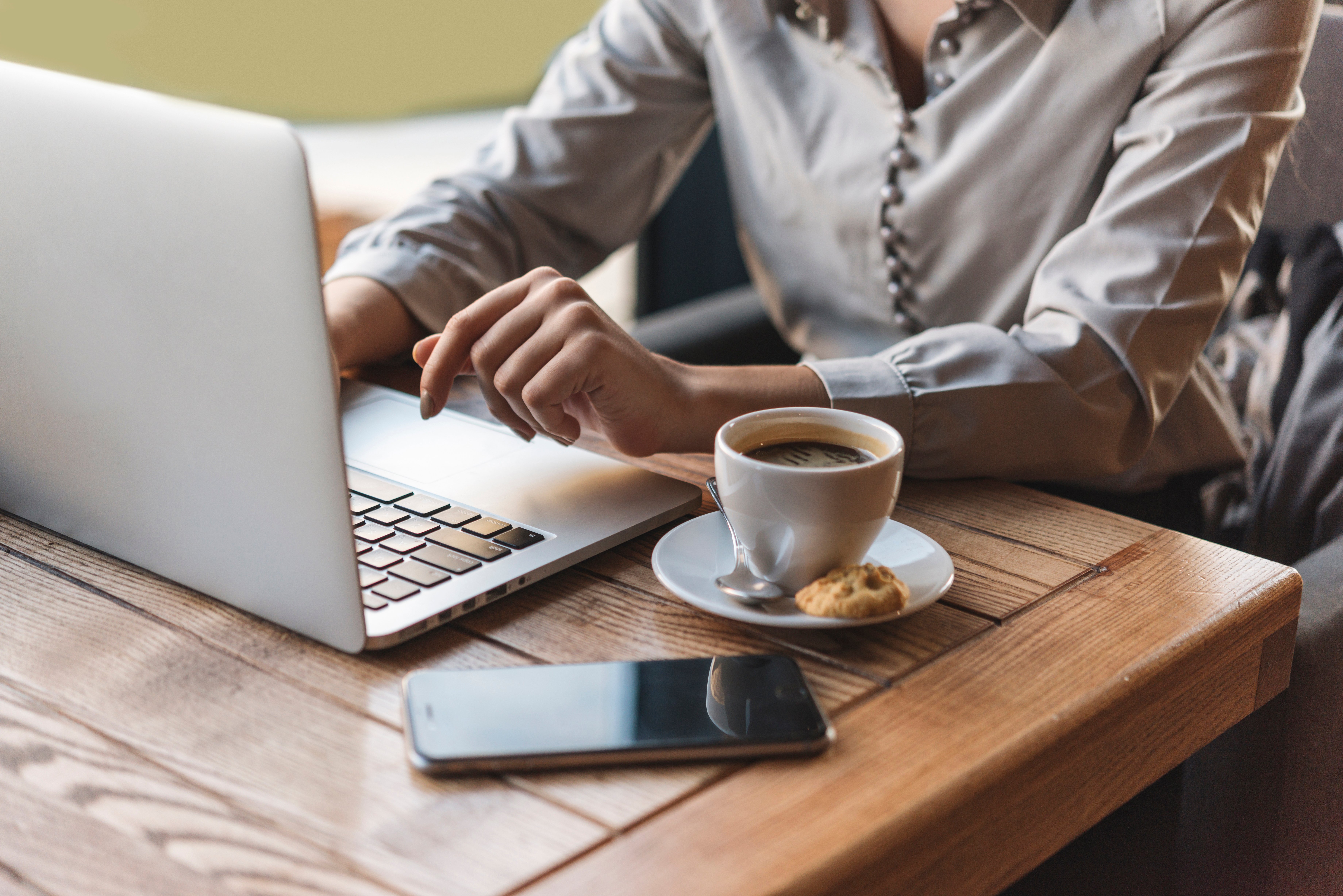 Persona en un ambiente de oficina acogedor trabajando en un computador portátil, con una taza de café y una galleta en la mesa de madera junto a su teléfono móvil